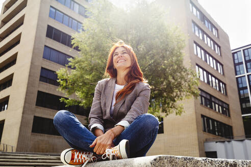 Lächelnde rothaarige Frau sitzt vor einem Gebäude an einem sonnigen Tag - IHF01150