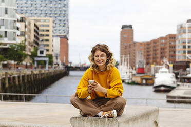 Happy woman with smart phone sitting on bench at Elbphilharmonie - IHF01158