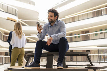 Smiling businessman using smart phone with colleagues in background at corridor - JOSEF12382