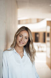 Smiling blond businesswoman leaning on wall in corridor - JOSEF12412