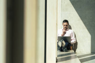 Businessman using laptop sitting on window sill in office - JOSEF12417