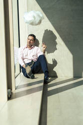 Thoughtful businessman sitting below artificial cloud looking through window at office - JOSEF12419