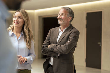 Happy businesswoman with arms crossed by colleague in corridor - JOSEF12471