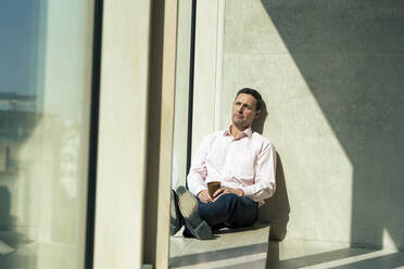 Contemplative businessman sitting on window sill in office - JOSEF12506