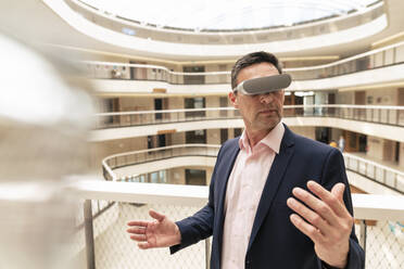 Mature businessman watching through VR glasses in corridor - JOSEF12513