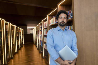 Professor mit Laptop am Bücherregal in der Bibliothek - JOSEF12586