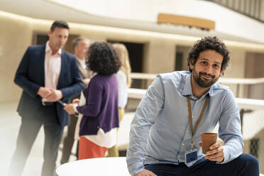 Lächelnder Geschäftsmann, der eine Kaffeetasse hält, während seine Kollegen im Hintergrund diskutieren - JOSEF12622