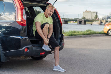 Reifer Mann bindet Schnürsenkel sitzend im Kofferraum eines Autos - VPIF07097