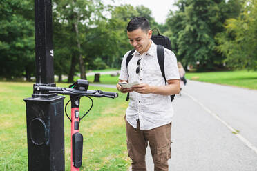 Man using smart phone standing near electric scooter at park - DMMF00001