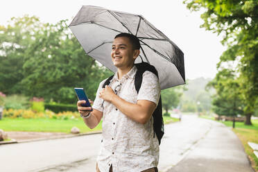Junger Mann mit Regenschirm und Smartphone im Park - DMMF00006