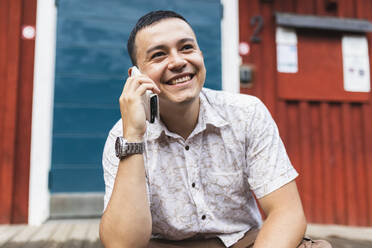 Glücklicher Mann, der mit einem Mobiltelefon spricht und vor einem Haus sitzt - DMMF00020