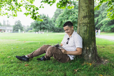 Smiling man reading book leaning on tree trunk at park - DMMF00021