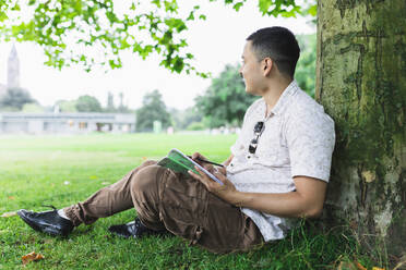 Young man with Sudoku book leaning on tree trunk at park - DMMF00022