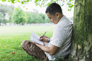 Man solving Sudoku in book sitting by tree trunk at park - DMMF00023