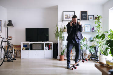 Cheerful businessman with skateboard talking on smart phone and clenching fist at home - ASGF02709