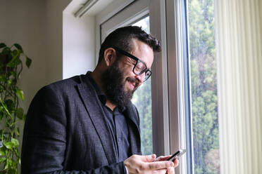 Lächelnder Freiberufler mit Brille, der sein Smartphone am Fenster zu Hause benutzt - ASGF02741