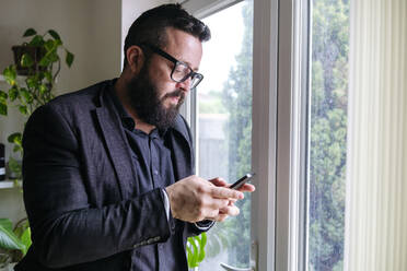 Älterer Geschäftsmann, der ein Mobiltelefon in der Nähe eines Fensters zu Hause benutzt - ASGF02743