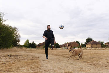 Mann spielt mit Hund, der einen Fußball auf einem Feld bei Sonnenuntergang kickt - ASGF02752