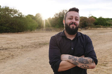 Smiling hipster man standing with arms crossed in field - ASGF02754