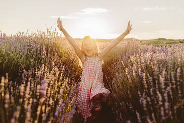 Fröhliches Mädchen mit erhobenen Armen genießt inmitten von Blumen in einem Lavendelfeld bei Sonnenuntergang - SIF00385