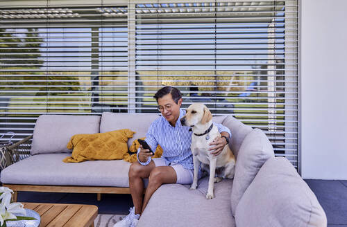 Lächelnder Mann, der ein Smartphone benutzt und mit seinem Hund auf dem Sofa sitzt - FMKF07654