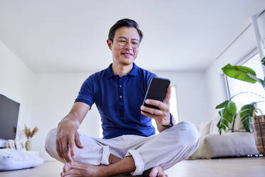Glücklicher reifer Mann, der mit seinem Mobiltelefon auf dem Fußboden zu Hause im Internet surft - FMKF07658