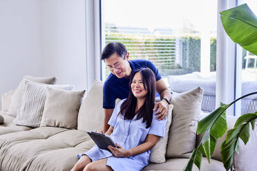 Happy man with woman holding tablet PC sitting on sofa at home - FMKF07703