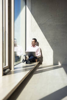 Geschäftsmann mit Laptop schaut durch ein Fenster im Büro - JOSEF12685