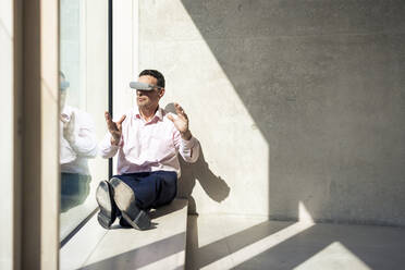 Businessman with VR glasses gesturing sitting on window sill in office - JOSEF12686