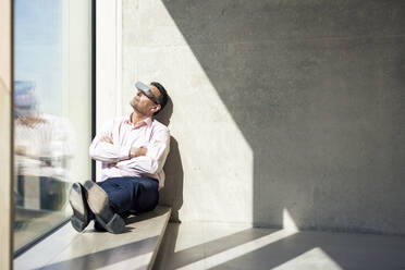 Businessman watching through smart glasses sitting on window sill in office - JOSEF12687