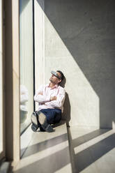Mature businessman watching through futuristic glasses sitting on window sill in office - JOSEF12688