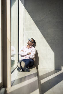 Älterer Geschäftsmann, der durch eine futuristische Brille auf der Fensterbank im Büro sitzt - JOSEF12688