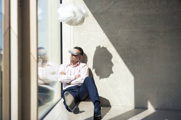 Businessman with arms crossed watching through smart glasses below artificial cloud in office - JOSEF12693