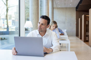 Kontemplativer Professor mit Laptop auf dem Schreibtisch in der Universität - JOSEF12766