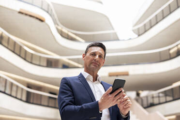 Confident mature businessman with smart phone in lobby - JOSEF12830