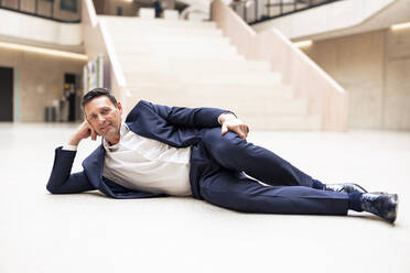 Smiling businessman lying on floor in lobby - JOSEF12833