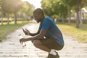 Happy man crouching on footpath using smart phone at park - EGAF02525