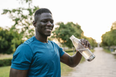 Smiling young man holding water bottle at park - EGAF02549