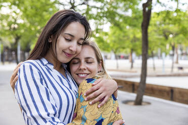 Smiling young women with eyes closed hugging each other at park - WPEF06297