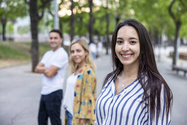 Smiling young woman with long hair standing in front of friends at park - WPEF06306