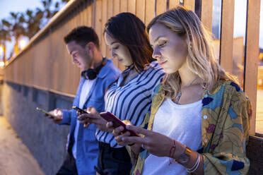 Junge Frauen und Mann surfen nachts vor einem Geländer mit ihren Smartphones im Internet - WPEF06321