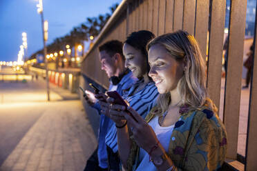 Smiling women and man text messaging through mobile phones in front of railing at night - WPEF06322