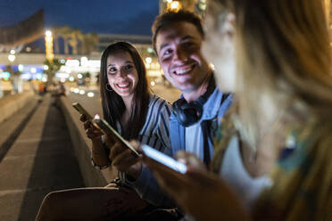Glücklicher junger Mann und Frau mit Handy in der Hand, die sich nachts mit einem Freund unterhalten - WPEF06330