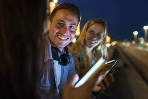 Fröhlicher junger Mann mit Smartphone inmitten von Freunden bei Nacht - WPEF06331