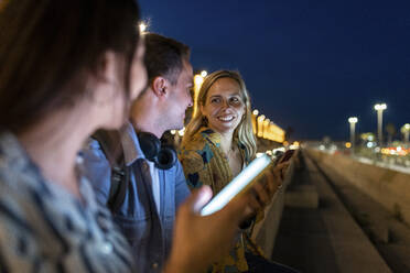 Glückliche Frau im Gespräch mit Freunden, die nachts ein Smartphone in der Hand halten - WPEF06332