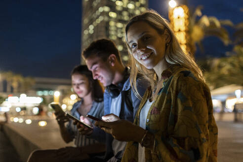 Lächelnde Frau mit Mobiltelefon, die nachts bei Freunden sitzt - WPEF06337