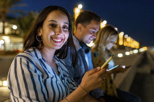 Glückliche junge Frau mit Smartphone, die abends bei Freunden sitzt - WPEF06338