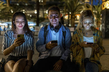 Lächelnder junger Mann sitzt inmitten von Frauen, die nachts ihr Smartphone benutzen - WPEF06339