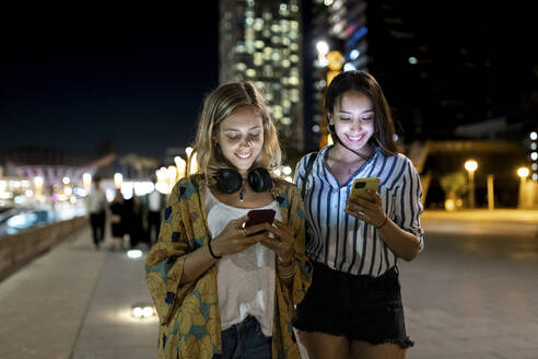 Lächelnde junge Frauen, die nachts in der Stadt ein Mobiltelefon benutzen - WPEF06350