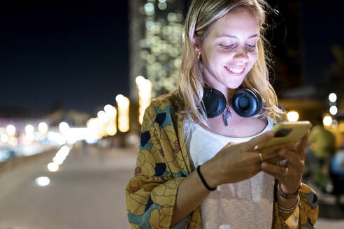 Glückliche Frau mit blondem Haar, die nachts ihr Smartphone benutzt - WPEF06356